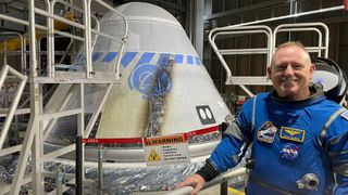 astronaut smiling next to a cone-shaped spaceship.  the spaceship is inside the launch gate and is surrounded by stairs.  the burn mark indicates the side of the spacecraft